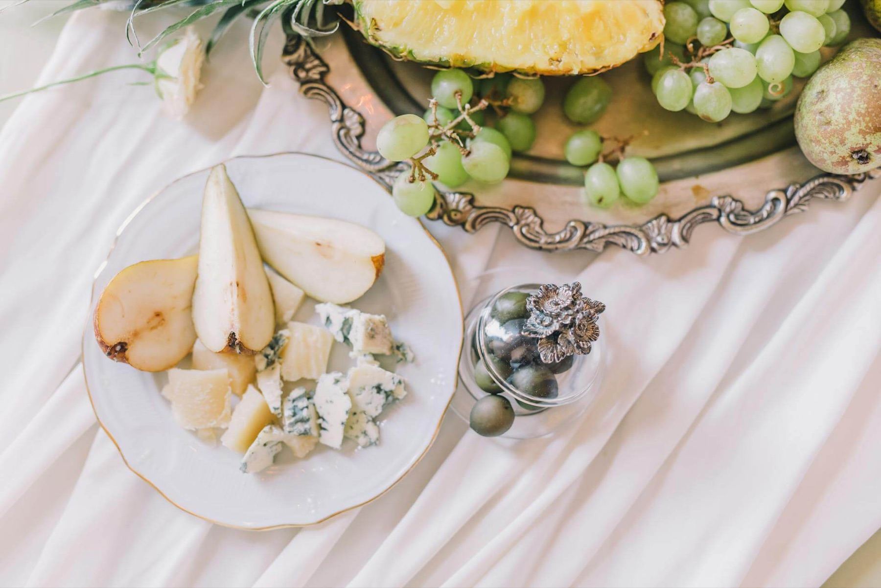 Vit tallrik med päronklyftor, lagrad ost och ädelost bredvid ett silverfat med gröna druvor och ananas på en vit draperad duk
