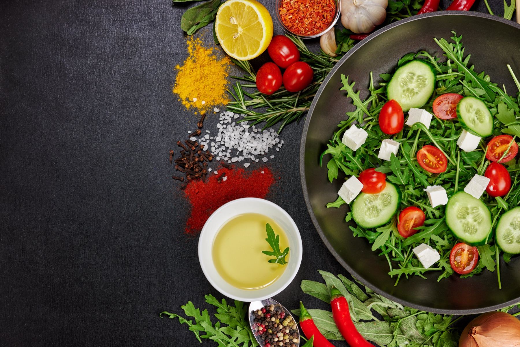 Svart panna med sallad av ruccola, gurka, tomat och fetaost, omgiven av citron, chili, färska örter och små högar kryddor på svart skiffer